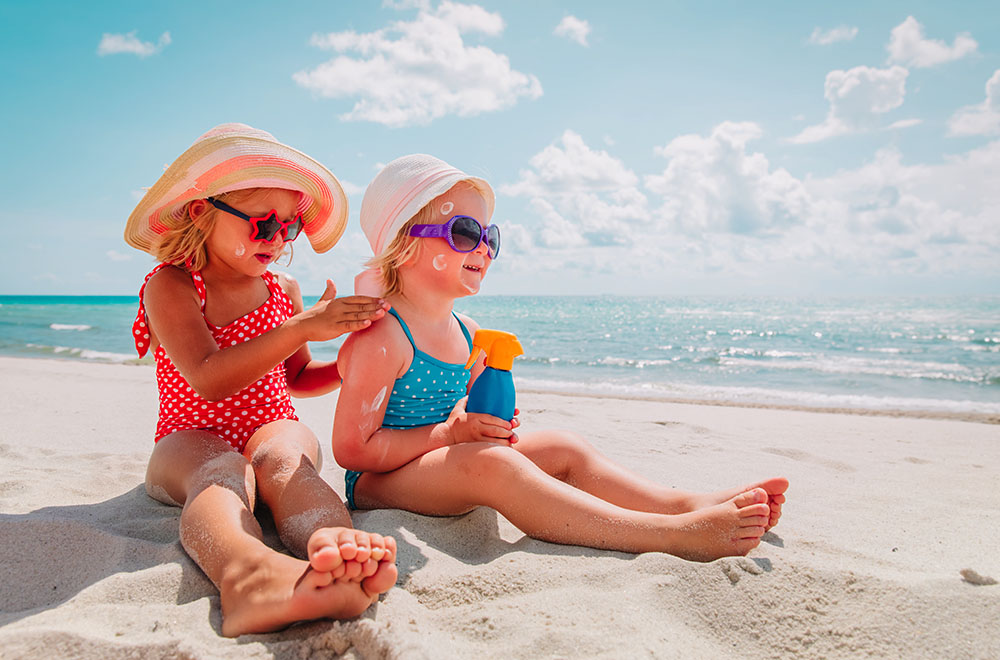 Dvije djevojke na plaži u kupaćim kostimima, nose naočale i šešire, nanose kremu za sunčanje