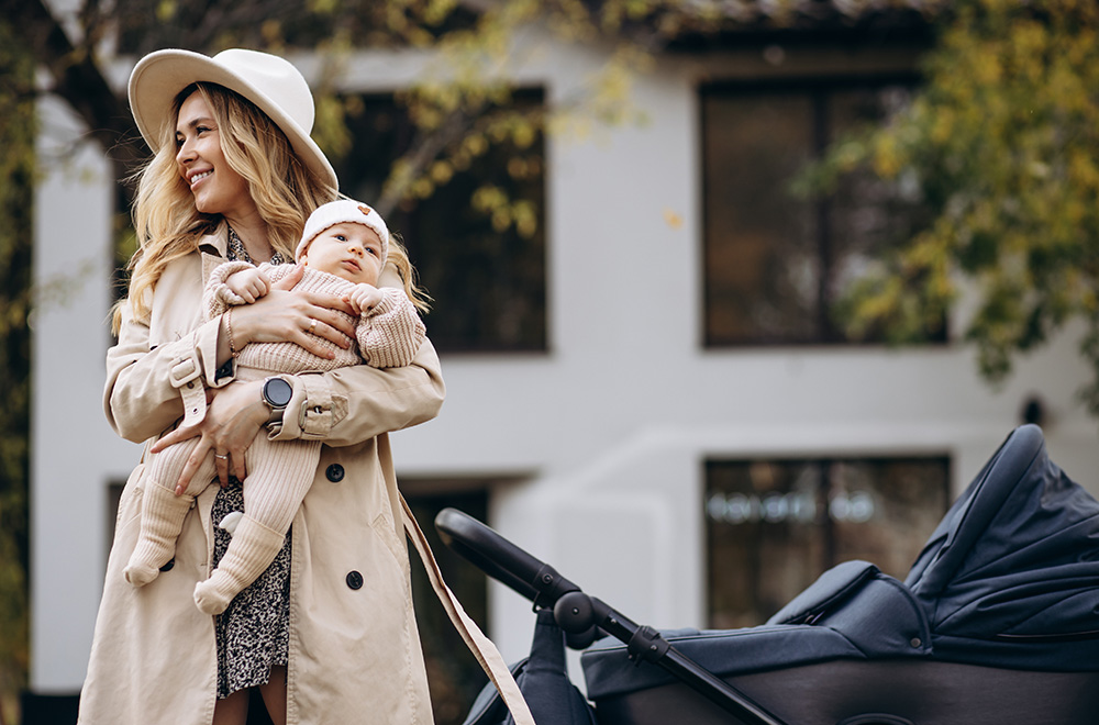 Žena drži bebu u naručju dok šeta pored kolica ispred zgrade.